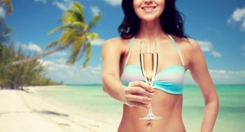 people, summer holidays, travel, celebration and drinks concept - happy young woman in bikini swimsuit drinking champagne at party over tropical beach background