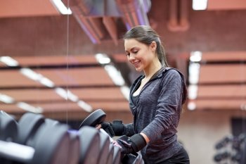 fitness, sport, exercising, weightlifting and people concept - young smiling woman choosing dumbbells in gym