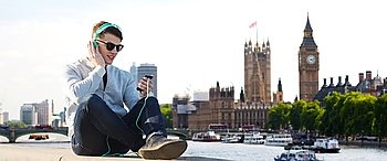 technology, travel, tourism and people concept - smiling young man or teenage boy in headphones with smartphone listening to music over london city and big ben tower background