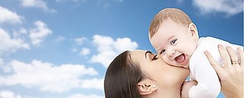 family, motherhood, parenthood, love and happy people concept - happy mother kissing her baby over blue sky and clouds background