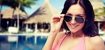 people, summer holidays, travel and tourism concept - happy young woman in sunglasses and pink swimsuit looking at you over hotel resort with swimming pool, bungalow and palm trees background