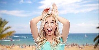 fun, expressions, summer holidays, travel and people concept - happy smiling young woman making bunny ears over exotic tropical beach with palm trees and sea background