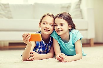 people, children, technology, friends and friendship concept - happy little girls lying on floor and taking selfie with smartphone at home