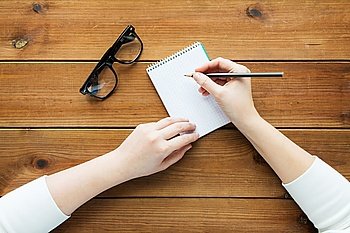 education, business and people concept - close up of woman or student writing to notepad with pencil and eyeglasses on wooden table