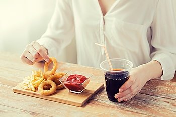 fast food, people and unhealthy eating concept - close up of woman eating deep-fried squid rings, french fries with ketchup and drinking cola on wooden table