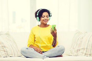 people, technology and leisure concept - happy african american young woman sitting on sofa with smartphone and headphones listening to music at home