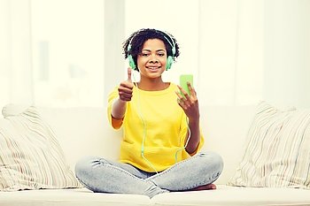 people, technology and leisure concept - happy african american young woman sitting on sofa with smartphone and headphones and showing thumbs up listening to music at home