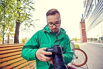 people, photography, technology, leisure and lifestyle - young hipster man holding and looking to digital camera with big lens on city street