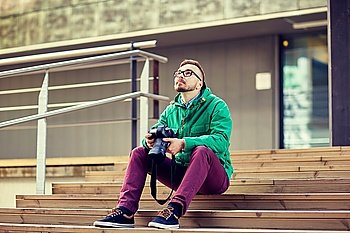 people, photography, technology, leisure and lifestyle - happy young hipster man holding digital camera with big lens and sitting on stairs in city