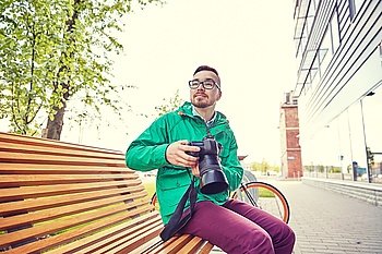people, photography, technology, leisure and lifestyle - happy young hipster man holding digital camera with big lens and looking for subject on city street