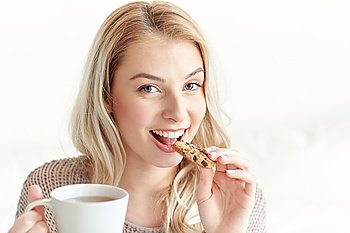 morning, breakfast, food and people concept - happy young woman with cup of tea eating cookie at home
