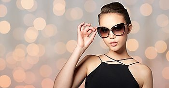 accessories, eyewear, fashion, people and luxury concept - beautiful young woman in elegant black sunglasses over holidays lights background