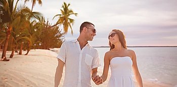 love, travel, summer holidays, people and relations concept - happy couple wearing sunglasses holding hands over exotic tropical beach with palm trees background