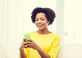 people, technology and leisure concept - happy african american young woman sitting on sofa with smartphone and earphones listening to music at home