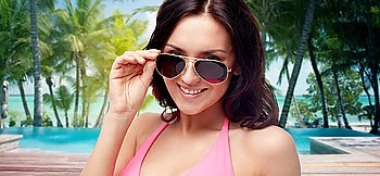 people, fashion, travel, tourism and summer concept - happy young woman in sunglasses looking at you over swimming pool and beach with palm trees background