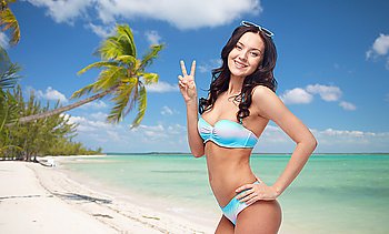 people, gesture, tourism, travel and summer concept - happy woman in bikini swimsuit showing victory hand sign over tropical beach background