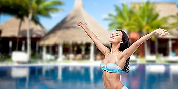 people, summer vacation, travel, tourism and freedom concept - happy woman in bikini swimsuit with raised hands looking up over swimming pool with bungalow and palm trees at hotel resort background
