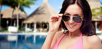 people, summer holidays, travel and tourism concept - happy young woman in sunglasses and pink swimsuit looking at you over hotel resort with swimming pool, bungalow and palm trees background