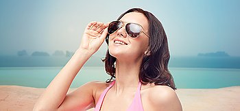 people, fashion, travel, tourism and summer concept - happy young woman in sunglasses and pink swimsuit looking up over infinity pool at sea side background