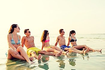 friendship, sea, summer vacation, holidays and people concept - group of smiling friends wearing swimwear and sunglasses sitting in water on beach
