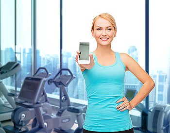 sport, fitness, technology, advertisement and people concept - smiling sporty woman showing smartphone blank screen over gym machines background