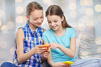 people, children, technology, friends and friendship concept - happy little girls with smartphones sitting on sofa at home over holidays lights background