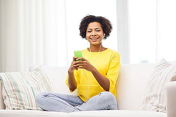 people, technology and leisure concept - happy african american young woman sitting on sofa with smartphone and earphones listening to music at home