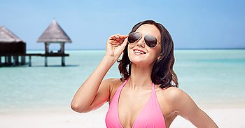 people, fashion, swimwear, summer and travel concept - happy young woman in sunglasses and pink bikini swimsuit over maldives beach with bungalow background