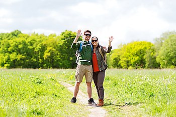 travel, hiking, backpacking, tourism and people concept - happy couple with backpacks waving hands and walking along country road outdoors