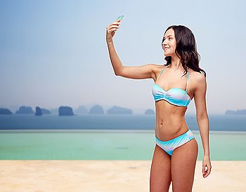 people, technology, summer holidays, travel and tourism concept - happy young woman in bikini swimsuit taking selfie with smatphone over beach or infinity edge pool and ocean background