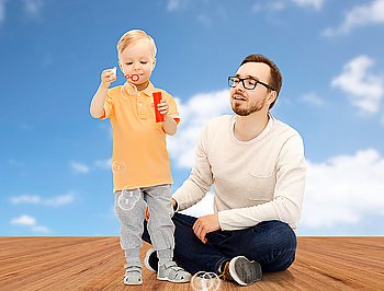 family, childhood, fatherhood, leisure and people concept - happy father and little son blowing bubbles and having fun over blue sky and wooden floor background