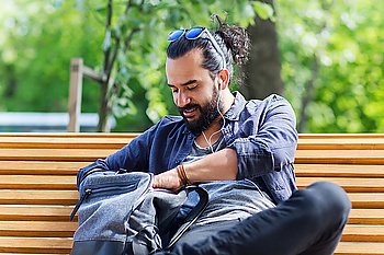 travel, tourism, lifestyle and people concept - man with earphones and sunglasses sitting on city bench and looking for something in his backpack