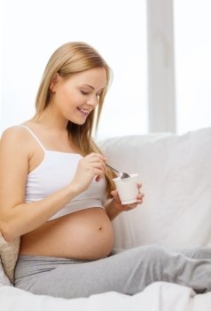 pregnancy, motherhood, healthcare, food and happiness concept - happy pregnant woman sitting on sofa with yogurt and spoon