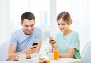 food, home, couple and technology concept - smiling couple with smartphones reading news and having breakfast at home