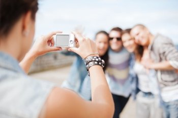 summer holidays and technology concept - close up of female hands holding digital camera and making photo of group of teenagers