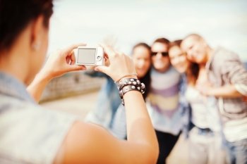 summer holidays and technology concept - close up of female hands holding digital camera and making photo of group of teenagers