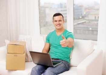 technology, home and lifestyle concept - smiling man with laptop and cardboard boxes at home showing thumbs up