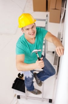 repair, building and home renovation concept - smiling man in yellow protective helmet hammering nail in wall