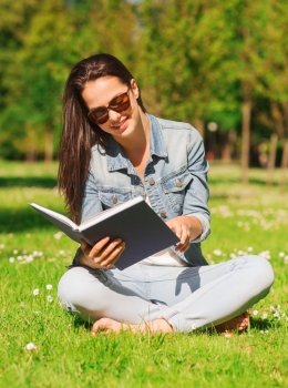 lifestyle, summer vacation, education, literature and people concept - smiling young girl reading book and sitting on grass in park