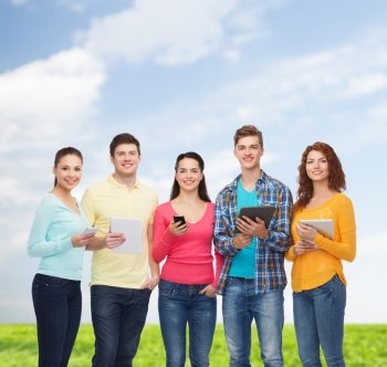 friendship, technology, summer and people concept - group of smiling teenagers with smartphones and tablet pc computers over blue sky and grass background