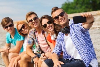 friendship, summer, technology and people concept - group of smiling friends with smartphone and headphones making selfie outdoors