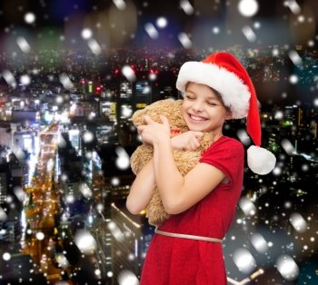 holidays, presents, christmas, childhood and people concept - smiling girl in santa helper hat with teddy bear over snowy night city background
