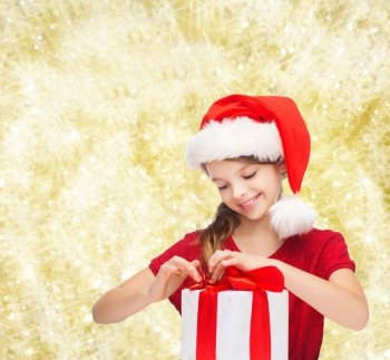 holidays, presents, christmas, childhood and people concept - smiling girl in santa helper hat with gift box over yellow lights background