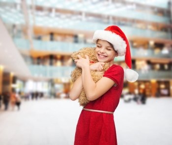 holidays, presents, christmas, childhood and people concept - smiling girl in santa helper hat with teddy bear over shopping center background