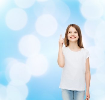 advertising, gesture, childhood and people - smiling little girl in white blank t-shirt pointing finger up over blue background