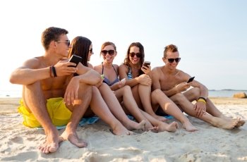 friendship, leisure, summer, technology and people concept - friends with smartphones sitting on sandy beach