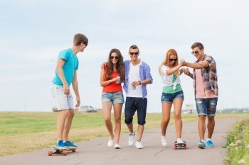 holidays, vacation, love and friendship concept - group of smiling teenagers walking and riding on skateboards outdoors