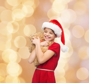 holidays, presents, christmas, childhood and people concept - smiling girl in santa helper hat with teddy bear over beige lights background