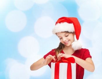 holidays, presents, christmas, childhood and people concept - smiling girl in santa helper hat with gift box over blue lights background