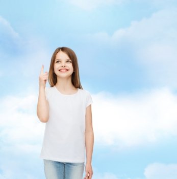 advertising, dream, childhood, gesture and people - smiling little girl in white blank t-shirt pointing finger up over cloudy sky background
